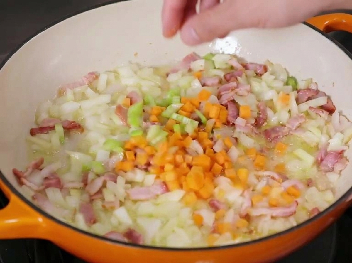 Gewürfelte Karotten werden in die Pfanne mit Zwiebeln, Sellerie und Speck gegeben.
