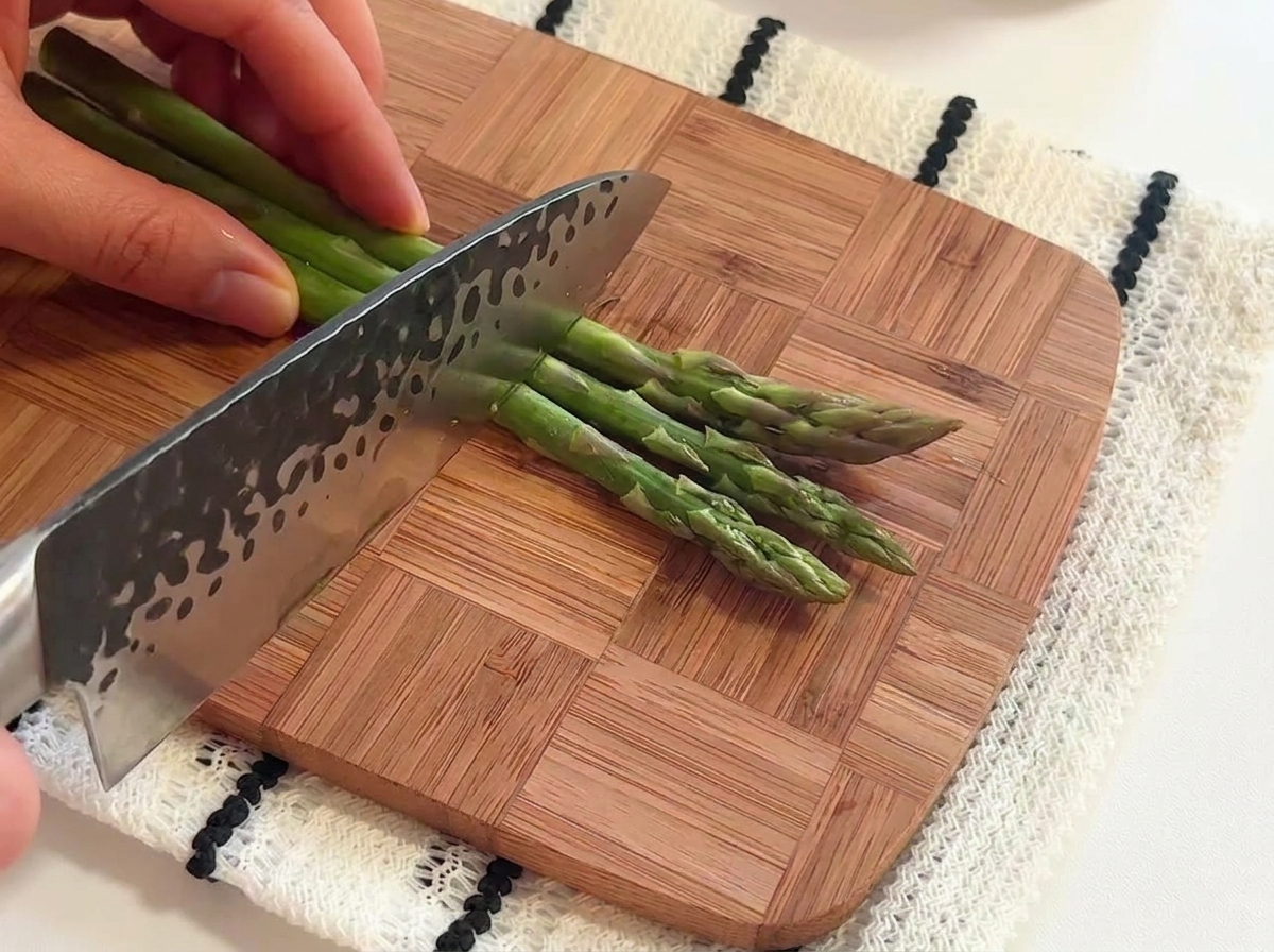 Hände schneiden frische grüne Spargelstangen mit einem Kochmesser auf einem Holzbrettchen in kurze Stücke.