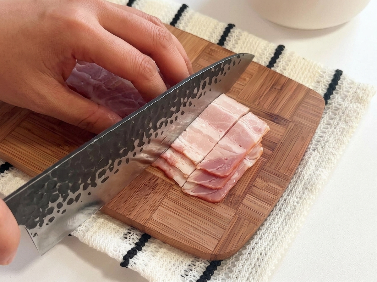 Rohe Speckstreifen werden mit einem großen Messer auf einem Holzbrett in kleine Stücke geschnitten.