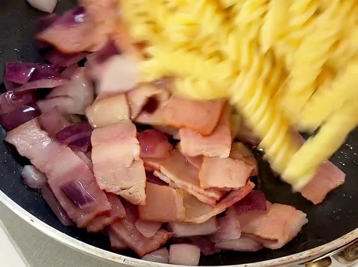 Gekochte Fusilli werden in die Pfanne zu Speck und Zwiebeln gegeben.