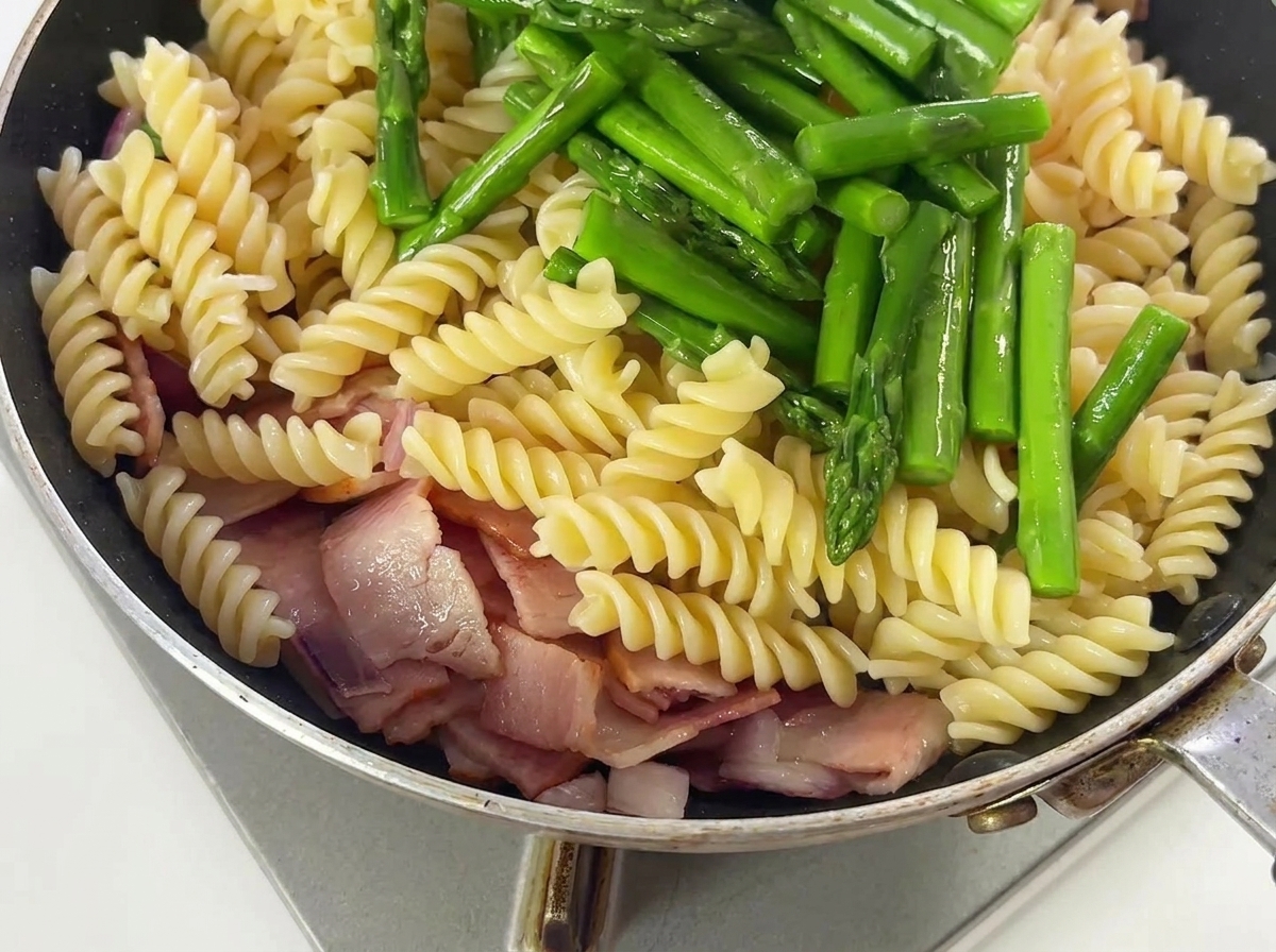 Spargelstücke und Gewürze werden mit Pasta, Speck und Zwiebeln in einer Pfanne geschwenkt.
