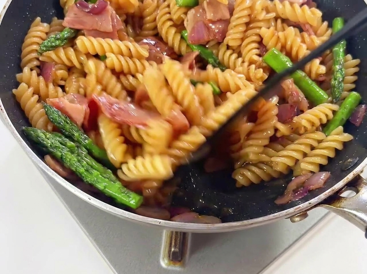 Pasta, grüner Spargel und Speckstücke werden in einer heißen Pfanne geschwenkt, um sich mit der Soße zu verbinden.