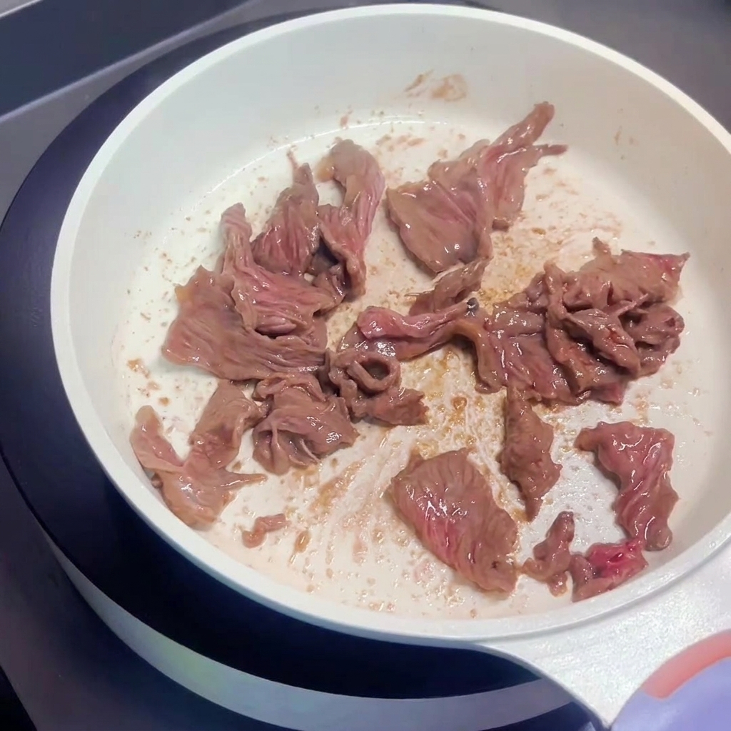 Rindfleischstreifen braten in der weißen Pfanne und zeigen braune Oberflächen.