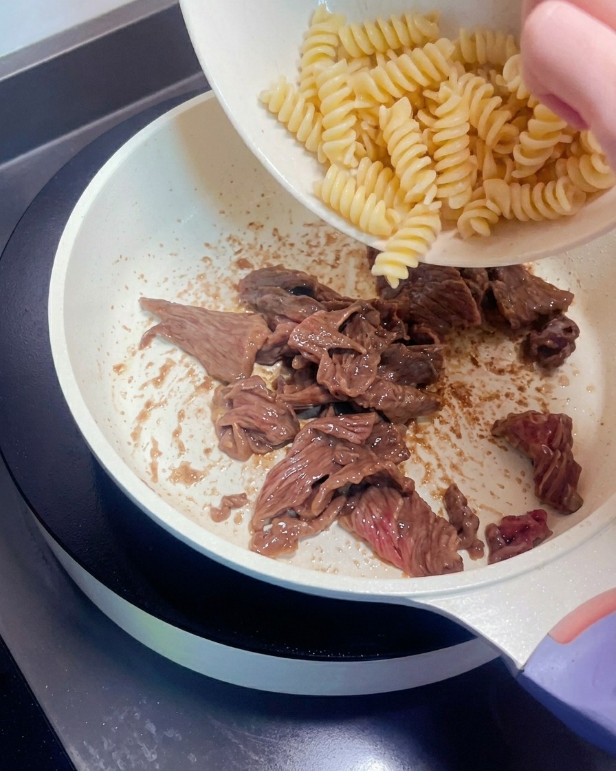 Spiralnudeln werden aus einer Schüssel zu dem gebratenen Fleisch in die Pfanne gegeben.