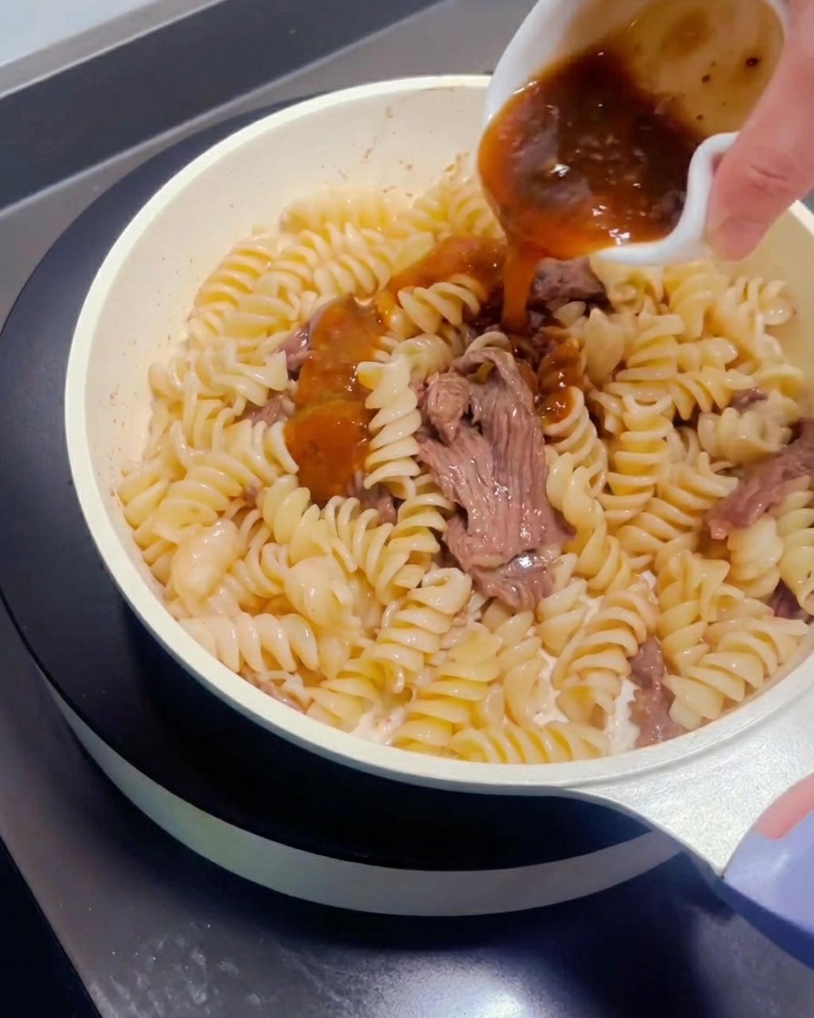 Pastasauce wird aus einem kleinen Schälchen über Fleisch und Nudeln in der Pfanne verteilt.