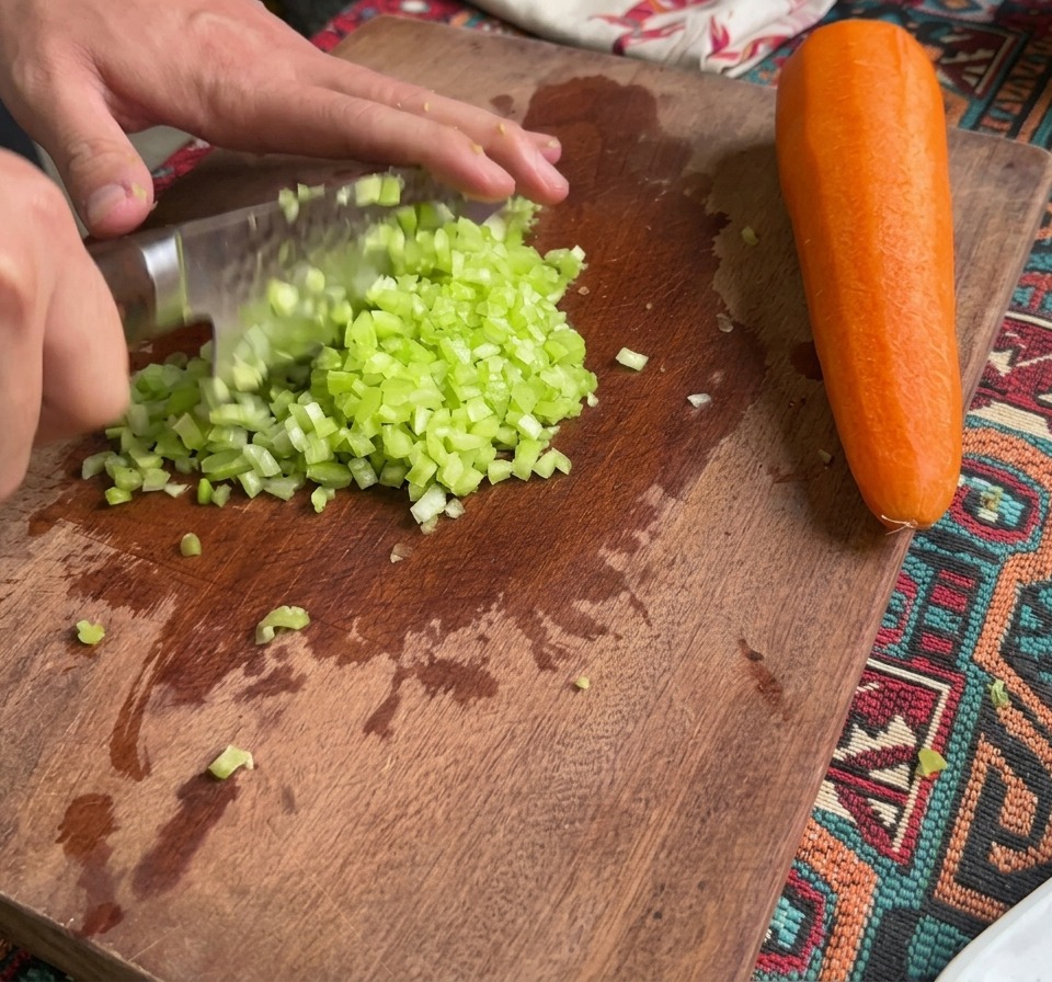 Fein gewürfelter Sellerie und Karotten liegen auf einem Holzschneidebrett neben einer ganzen Karotte.