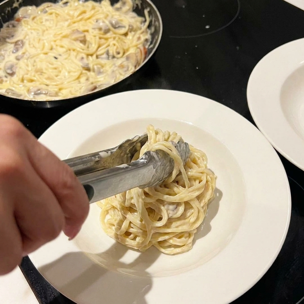 Cremige Nudeln werden in einer Pfanne geschwenkt, darüber wird Käse und Petersilie gestreut.