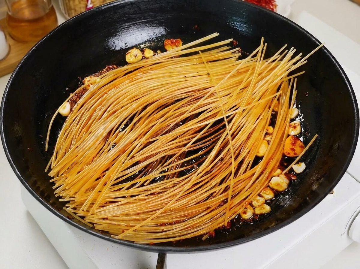 Eine Schöpfkelle gießt rote Tomatenbrühe über die angebratenen Spaghetti in einer schwarzen Pfanne.