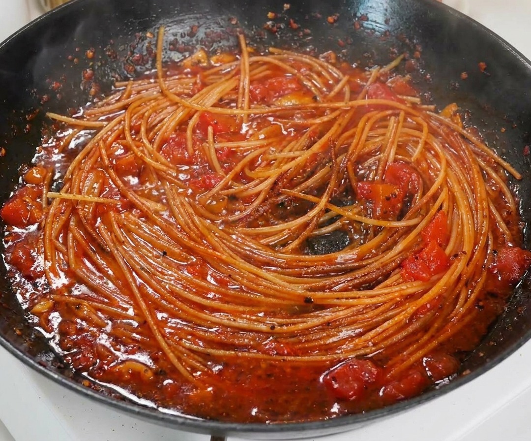 Nahaufnahme von gekochten Spaghetti, die in einer reichhaltigen, dicken Tomatensoße in einer schwarzen Pfanne geschwenkt werden.
