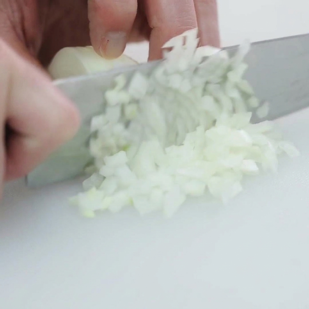 Eine weiße Zwiebel wird auf einem sauberen weißen Schneidebrett in kleine, gleichmäßige Stücke gewürfelt.