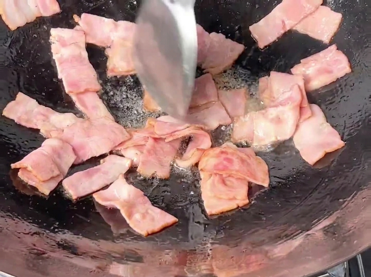 Rechteckige Speckstücke werden in einem dunklen Eisenwok mit einem Metallspatel angebraten.