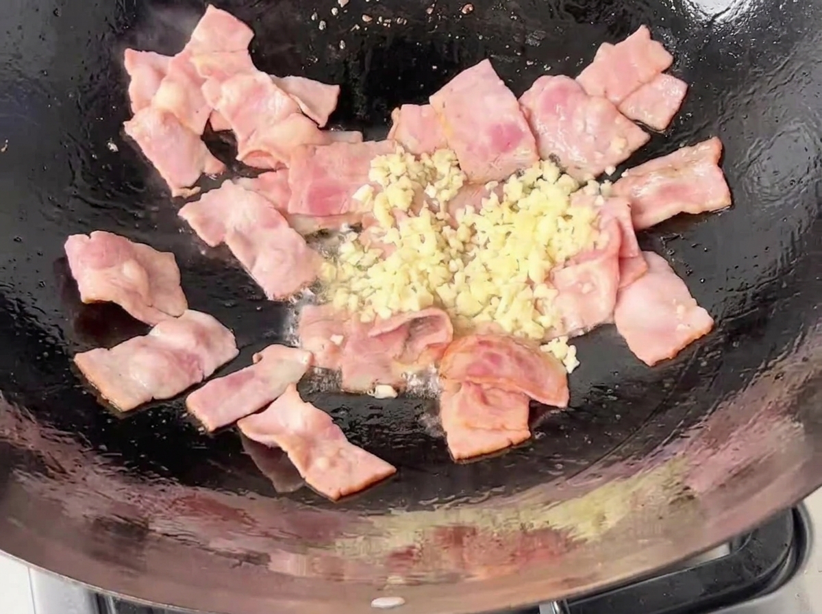 Fein gehackter weißer Knoblauch wird zu den brutzelnden, teilweise knusprigen Speckstücken in die Pfanne gegeben.