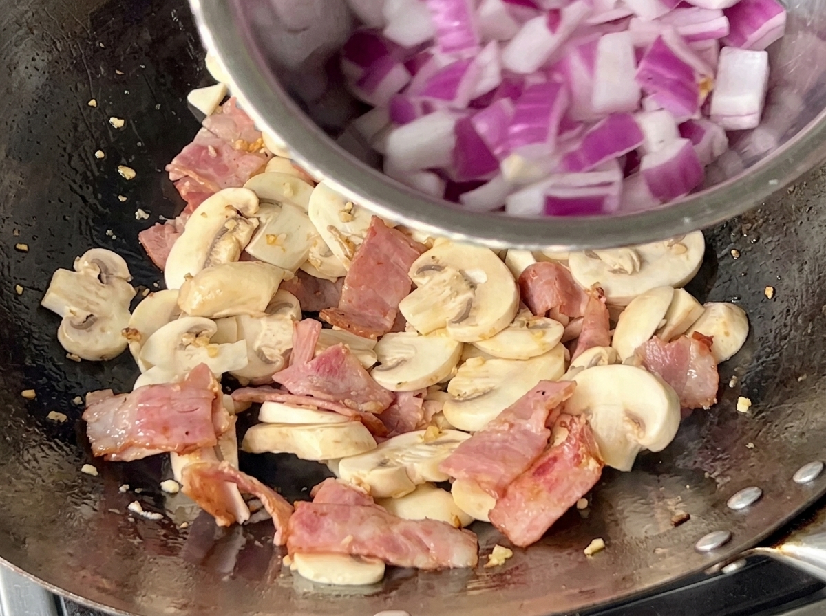 Ein Wok gefüllt mit geschnittenen Champignons, Speckwürfeln und frisch hinzugefügten gewürfelten roten Zwiebeln, die zusammen angebraten werden.
