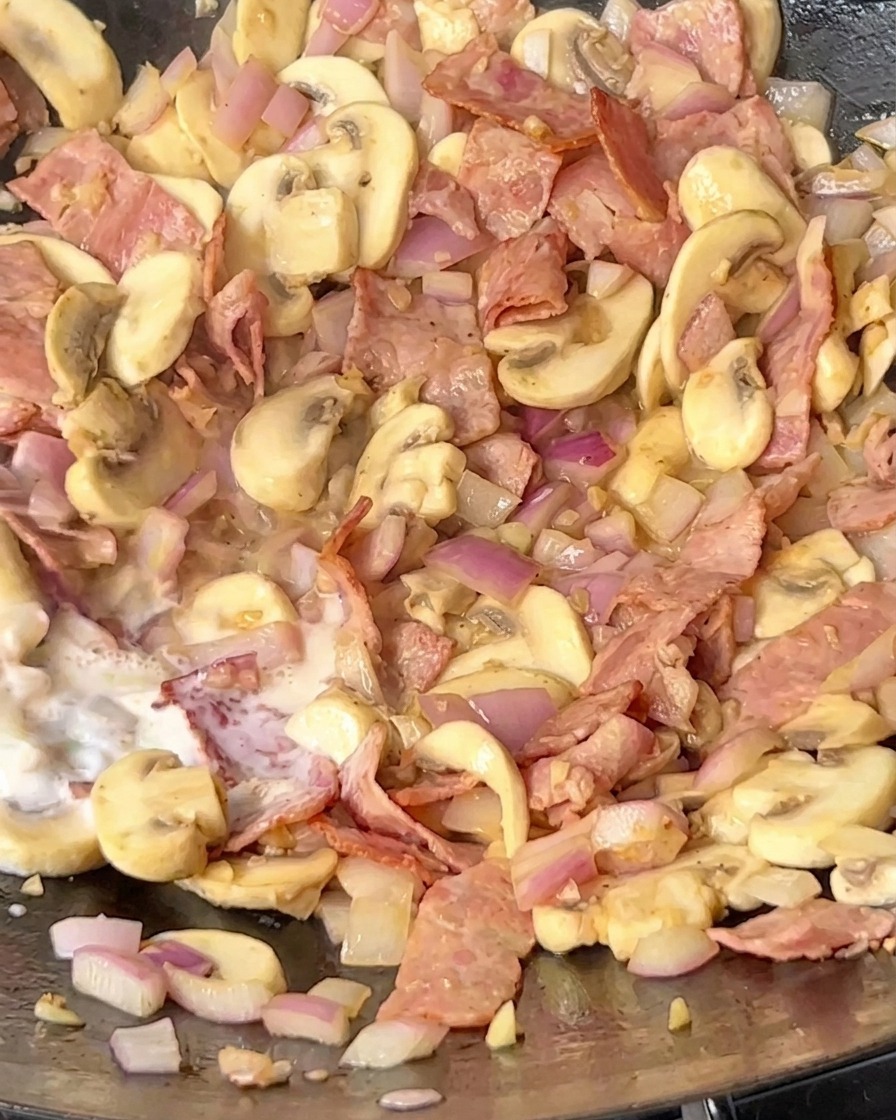 Milch wird in einen Wok gegossen, der angebratenen Speck, sautierte Pilze und Zwiebeln enthält.