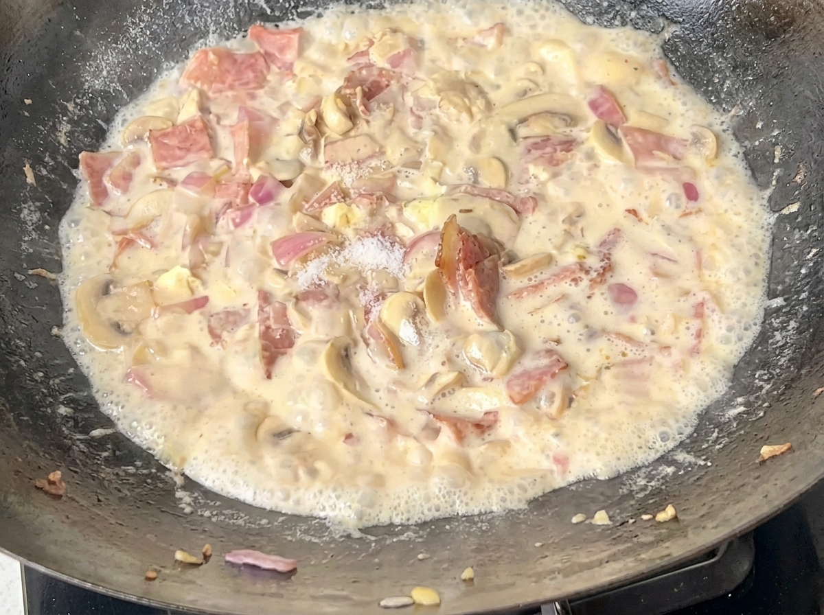 Gekochte Spaghetti werden in einen Wok gegeben, der mit cremiger Speck-Pilz-Sauce gefüllt ist.
