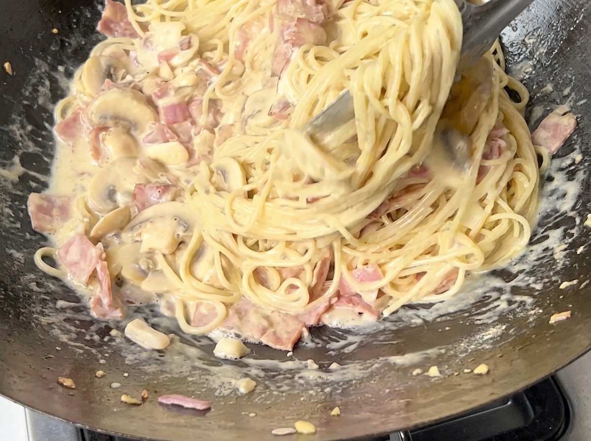 Eine Nahaufnahme von Spaghetti, die mit einer Metallzange in einer cremigen Speck-Pilz-Sauce geschwenkt werden.