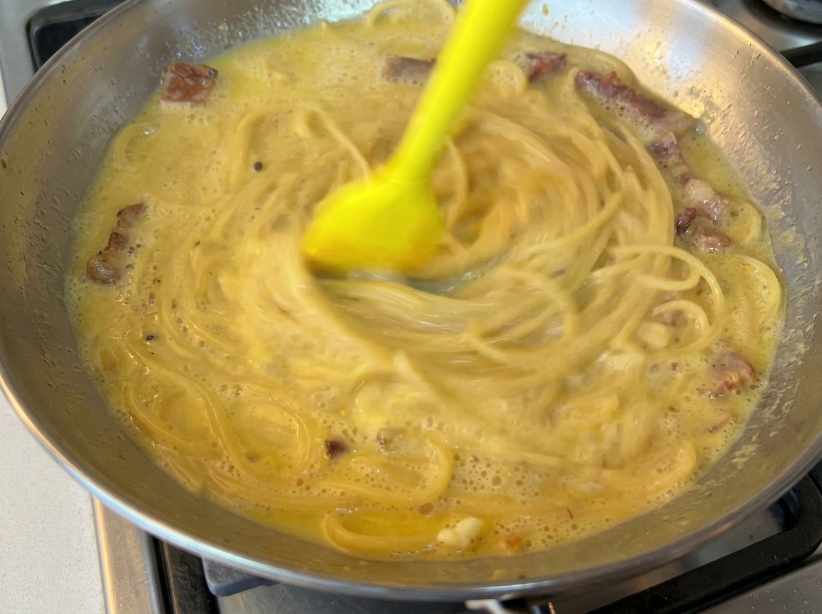 Die Nudeln werden mit einem Teigschaber in der Pfanne zur cremigen Sauce verrührt.