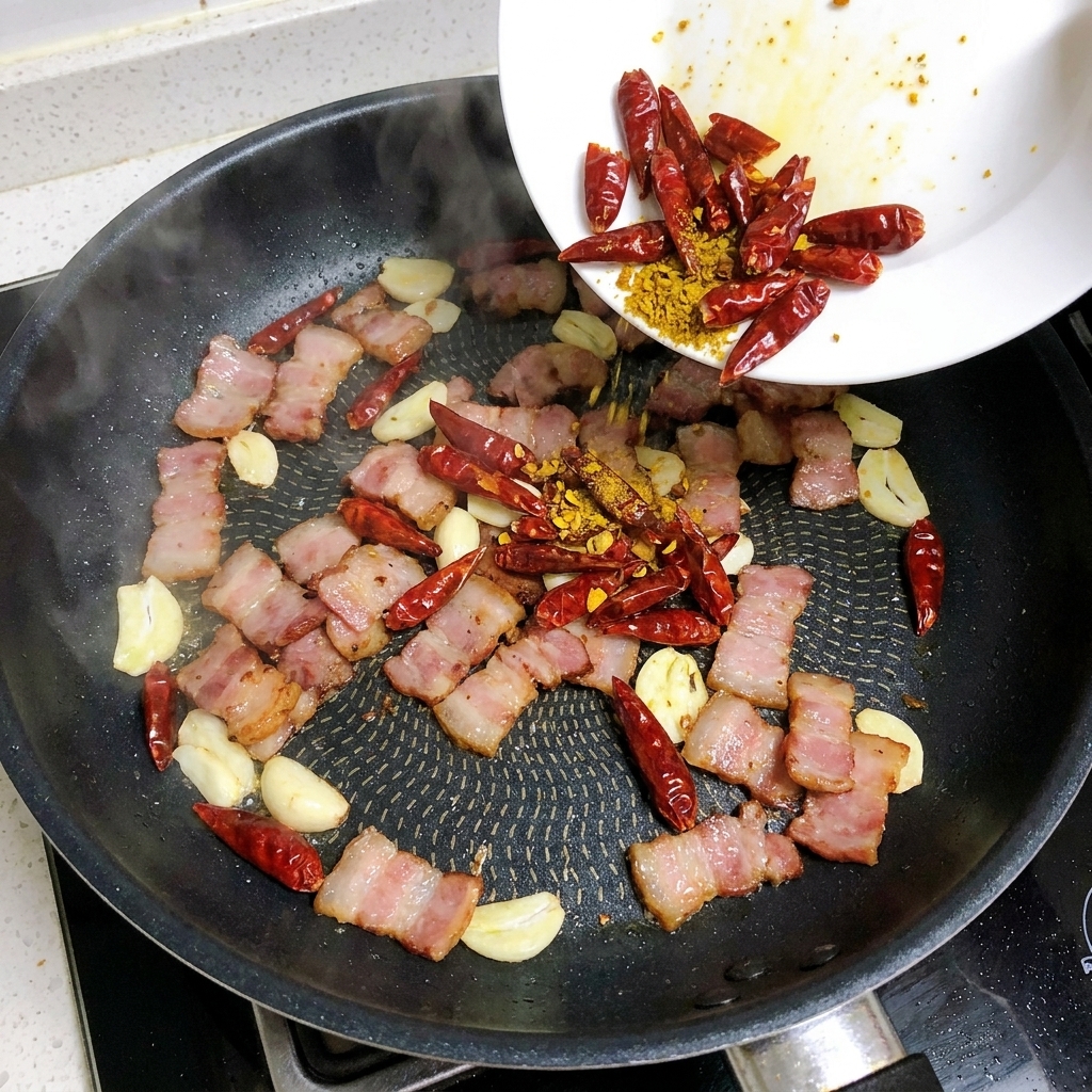 Knoblauchscheiben und getrocknete Chilis werden in die Pfanne mit dem gebratenen Speck gegeben.