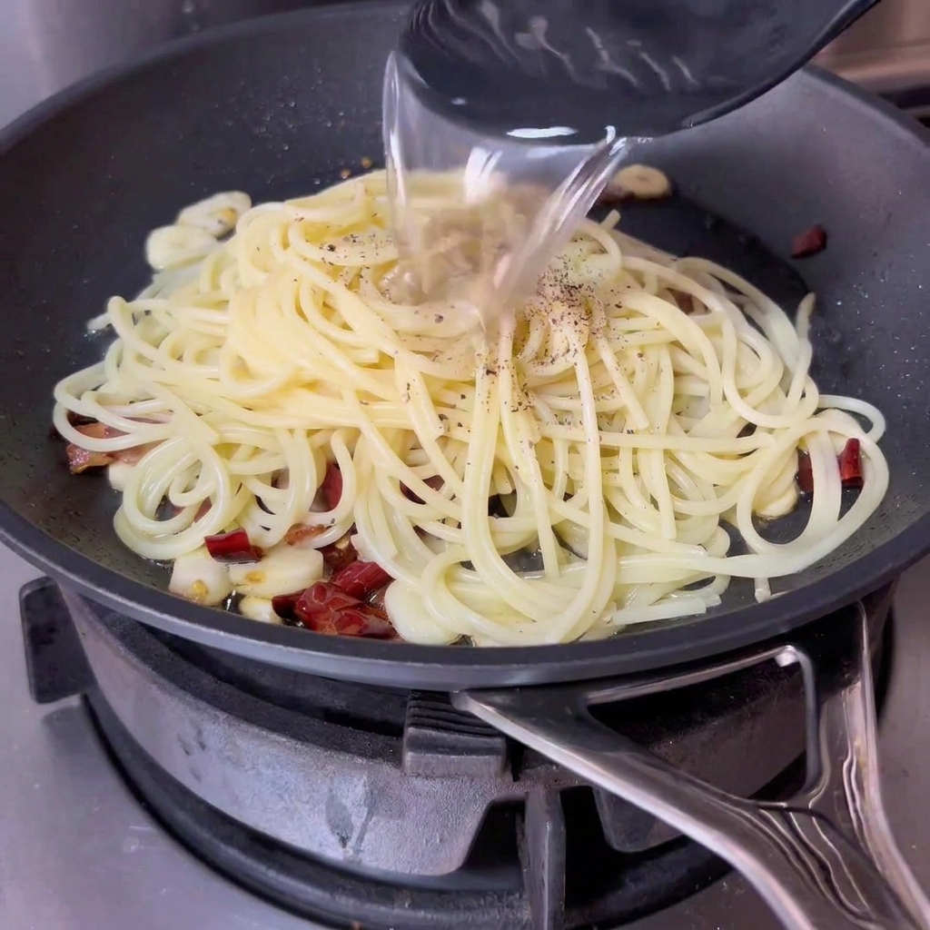 Eine Kelle gibt stärkehaltiges Nudelwasser in die Pfanne mit den Spaghetti und Speck.