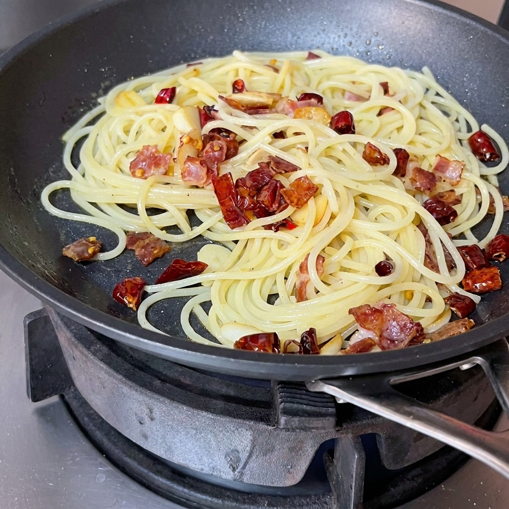 Spaghetti, Speck und Gewürze werden mit einem roten Pfannenwender in der Pfanne geschwenkt.