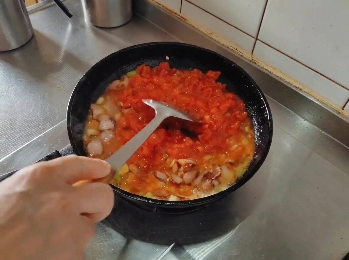 Ein Metallwender zerdrückt die Tomatenstücke in einer Pfanne mit Speck und Aromaten.