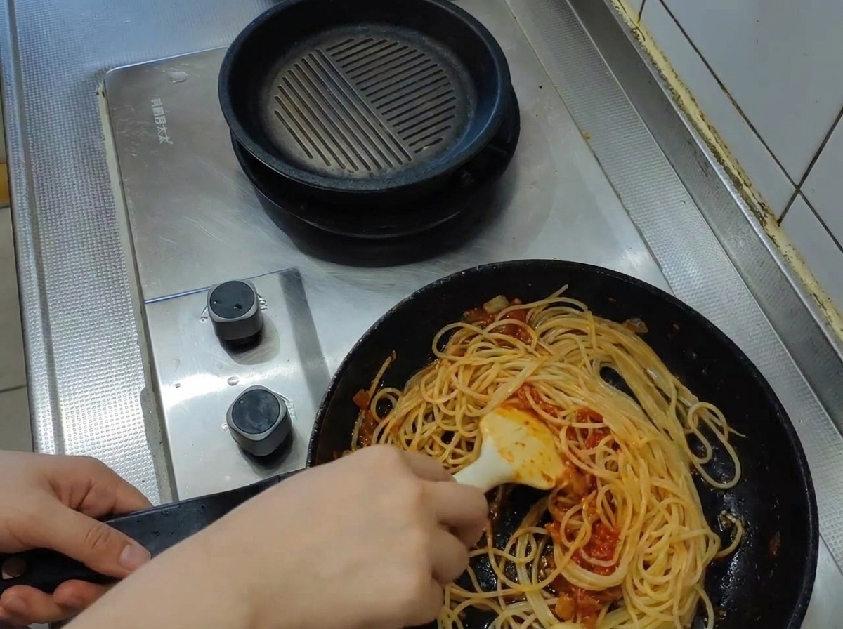 Eine Schöpfkelle gibt trübes, kochendes Nudelwasser in die Pfanne zu Pasta und Sauce.