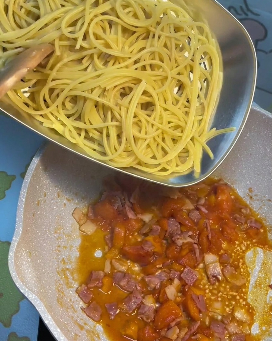 Gekochte Pasta wird direkt in die Pfanne mit der sprudelnden Tomaten-Speck-Sauce gegeben.