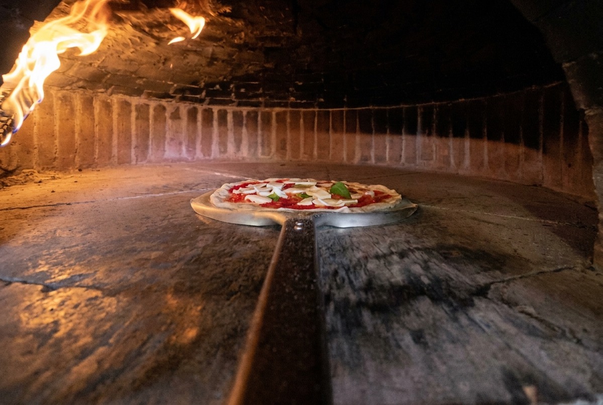 Eine Metallschaufel schiebt die rohe Pizza in einen heißen Holzofen.