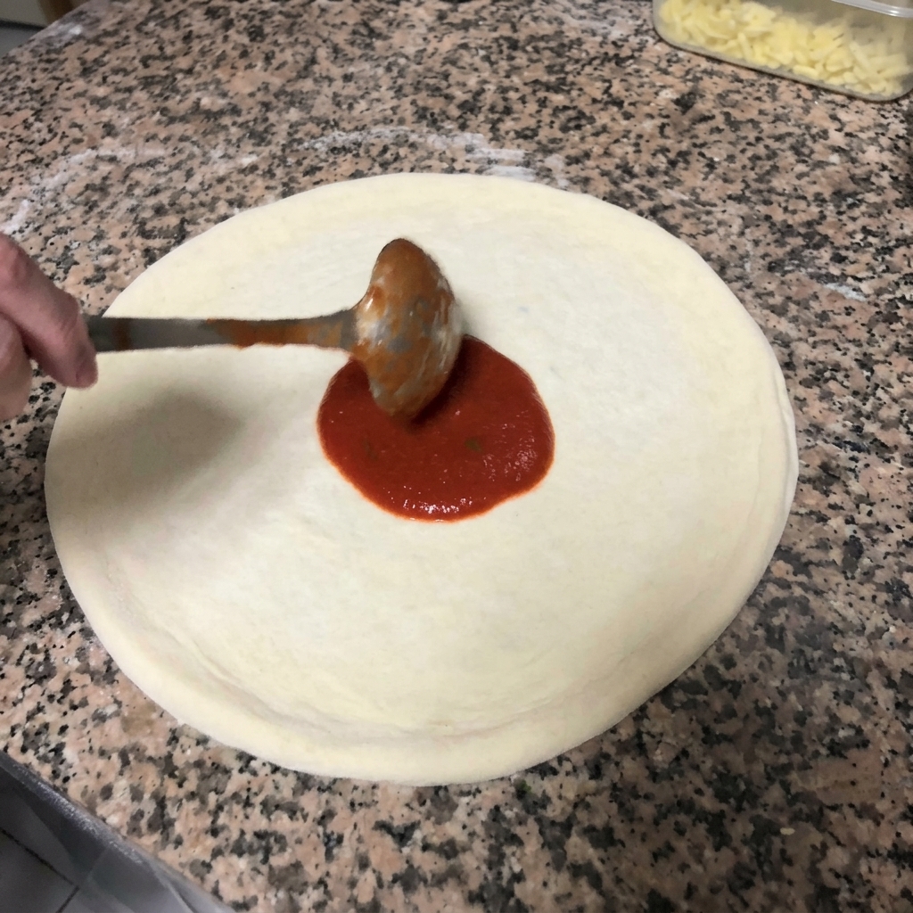 Die rote Tomatensauce wird mit einer Kelle in kreisenden Bewegungen auf dem Pizzateig verteilt.