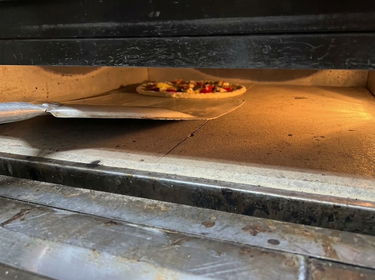 Eine Pizza wird mit einem Pizzaschieber in einen heißen Ofen auf einen Backstein geschoben.