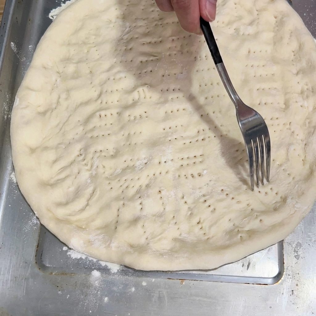Eine Gabel sticht den flachen Teigboden auf dem Backblech ein.