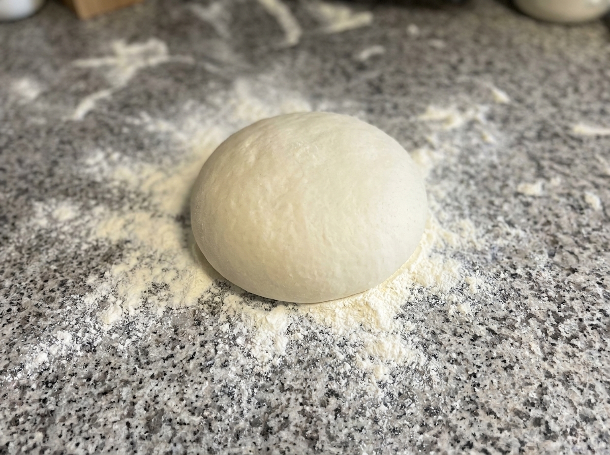 Teig, der auf einer bemehlten Arbeitsfläche zu einem Kreis geformt wird.