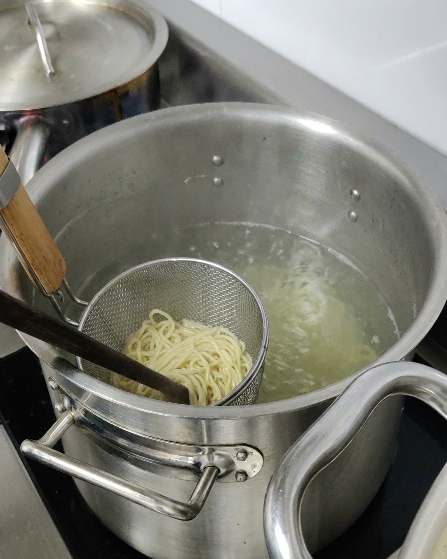 Ramen-Nudeln kochen in einem Metallsieb, das in einen großen Topf mit heißem Wasser getaucht ist.