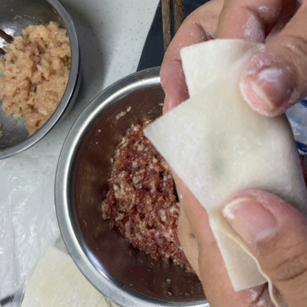 Hände falten ein Wonton-Teigblatt um die Füllung und verschließen die Ränder mit Wasser.