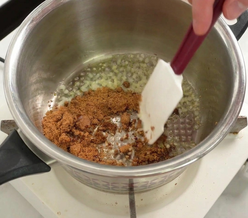 Brauner Zucker wird mit einem Spatel in den Topf zu den Zwiebeln und dem Knoblauch gerührt.
