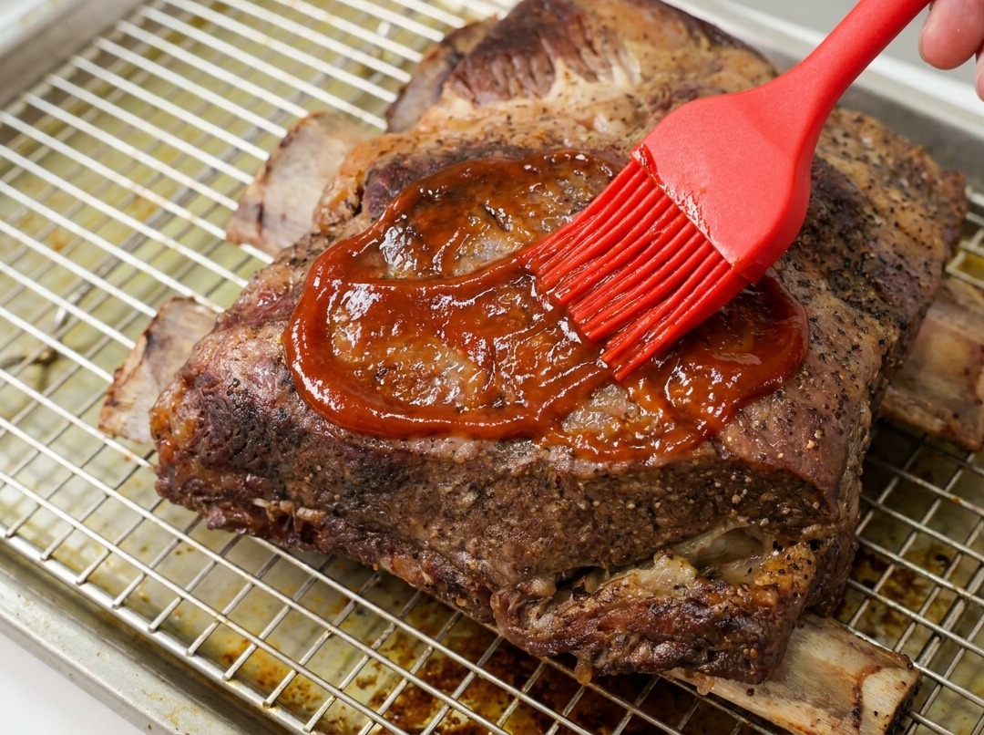 Ein Silikonpinsel verteilt glänzende BBQ-Sauce auf den gegarten Rinderrippen.