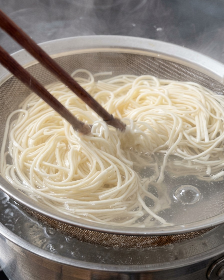 Frische Ramen-Nudeln werden mit Holzstäbchen aus kochendem Wasser in einem Metallsieb angehoben.