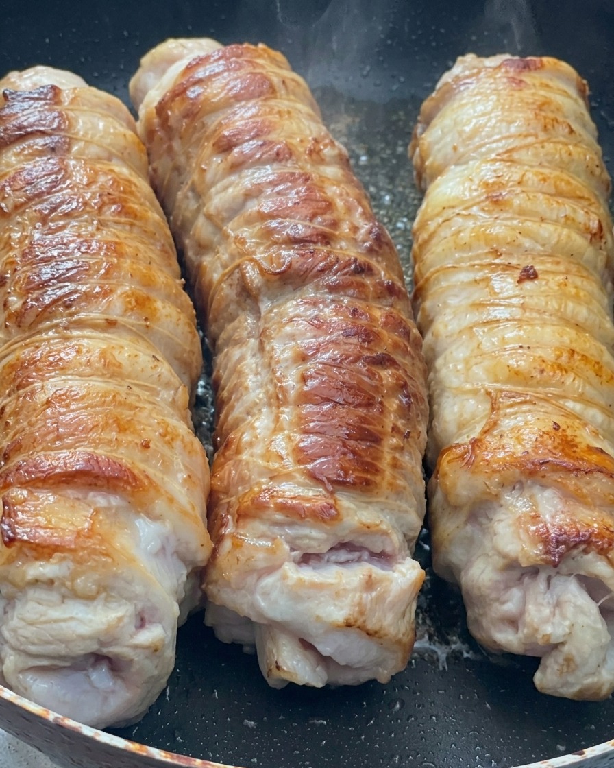 Drei große gerollte Stücke Schweinebauch braten in einer heißen Pfanne und entwickeln eine goldbraune Kruste.