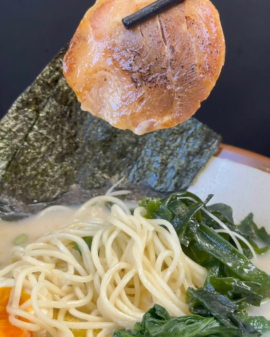 Schwarze Stäbchen heben eine Scheibe karamellisiertes Chashu über eine Schüssel mit Ramen, Nudeln und Toppings.