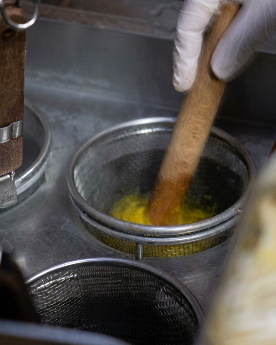Ein Metall-Nudelsieb mit gelben Nudeln wird mit einem Holzlöffel in einem Topf mit sprudelnd kochendem Wasser gerührt.
