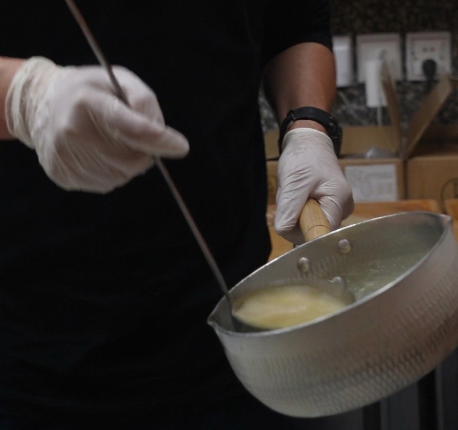 Handschuhe halten einen Metalltopf und eine Schöpfkelle und gießen cremige gelbe Chicken Paitan Brühe ein.