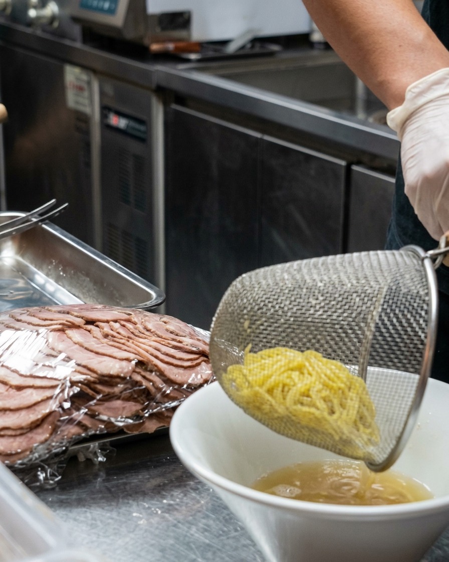 Ein Metallsieb lässt frisch gekochte gelbe Ramen-Nudeln in eine weiße Schüssel mit cremiger Brühe fallen.