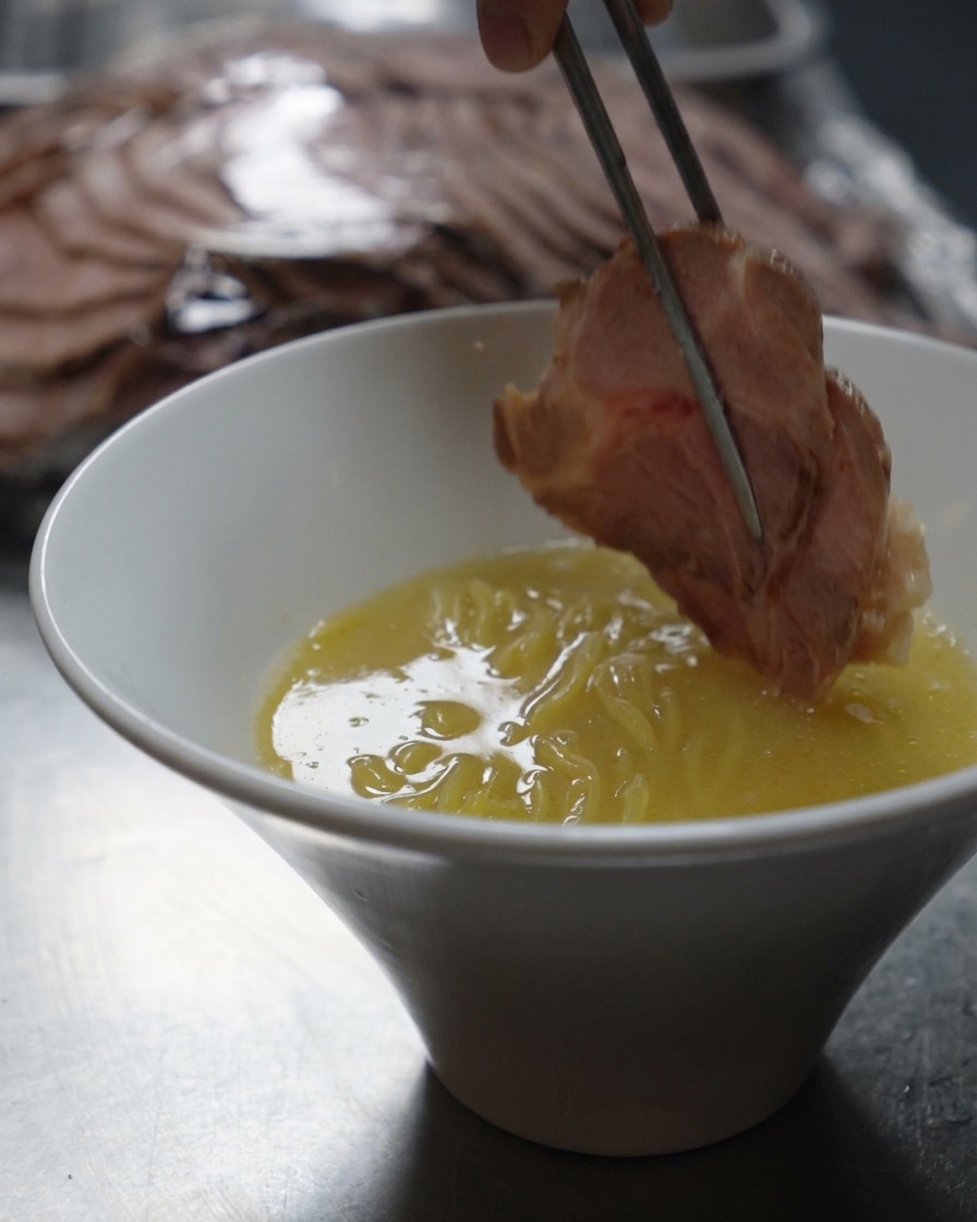 Metallstäbchen legen eine dicke Scheibe gebratenes Schweinefleisch in eine Schüssel mit Ramen-Nudeln und cremiger Brühe.