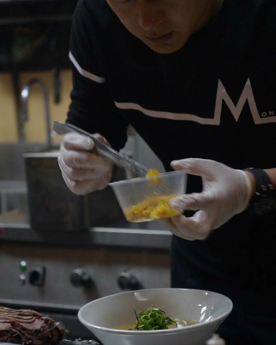 Ein kleiner Löffel mit leuchtend gelber Yuzu-Zeste wird über die fertige Ramen gestreut.