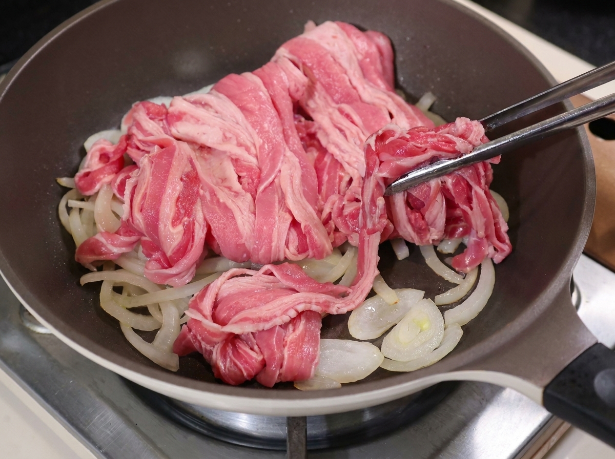 Zange legt dünne Rindfleischscheiben auf die Zwiebeln in der heißen Pfanne.