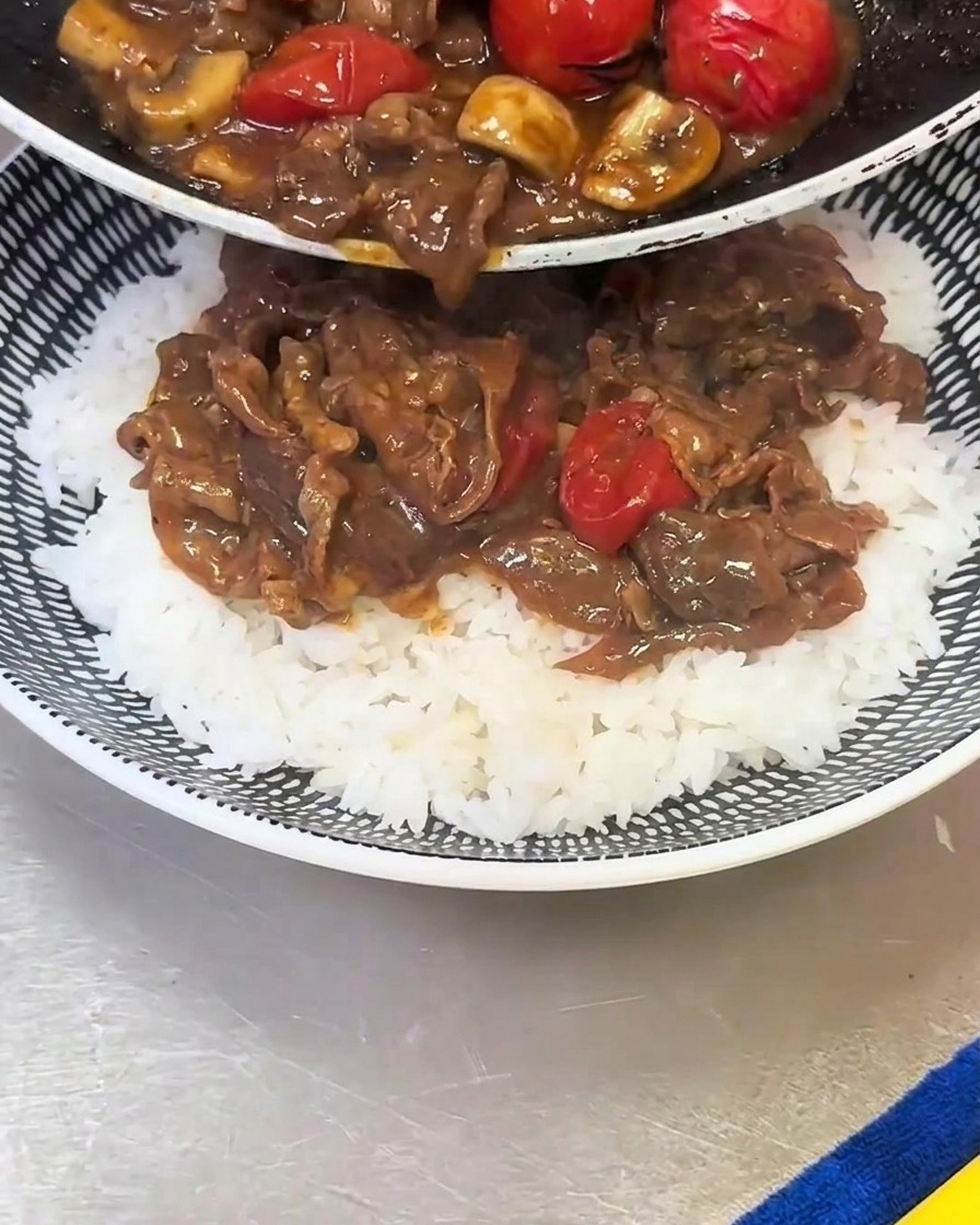 Die gekochte Mischung aus Rindfleisch, Pilzen und Kirschtomaten wird aus einer Pfanne auf eine Schüssel mit weißem Reis gegossen.