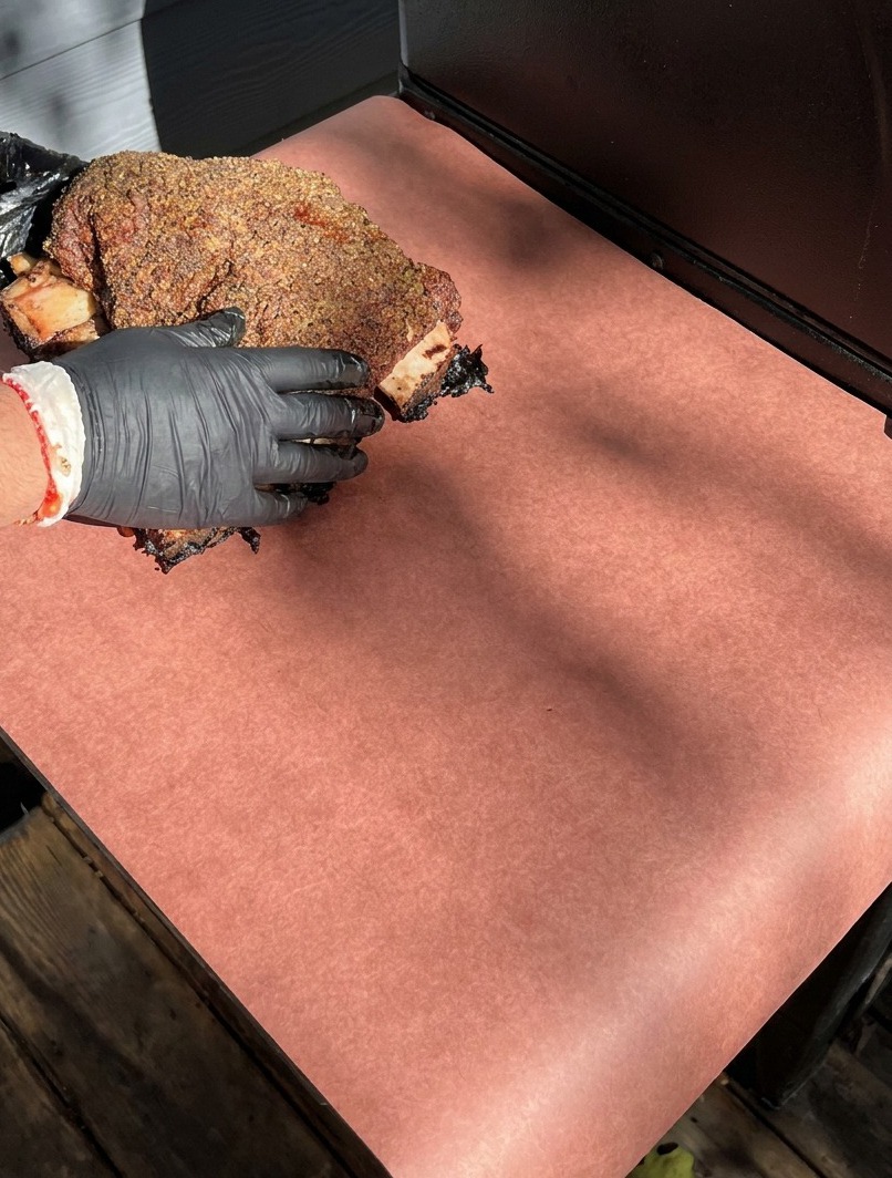 Hände in schwarzen Handschuhen legen dunkle, geräucherte Rinderrippen auf ein großes Blatt rosa Butcher Paper zum Einwickeln.