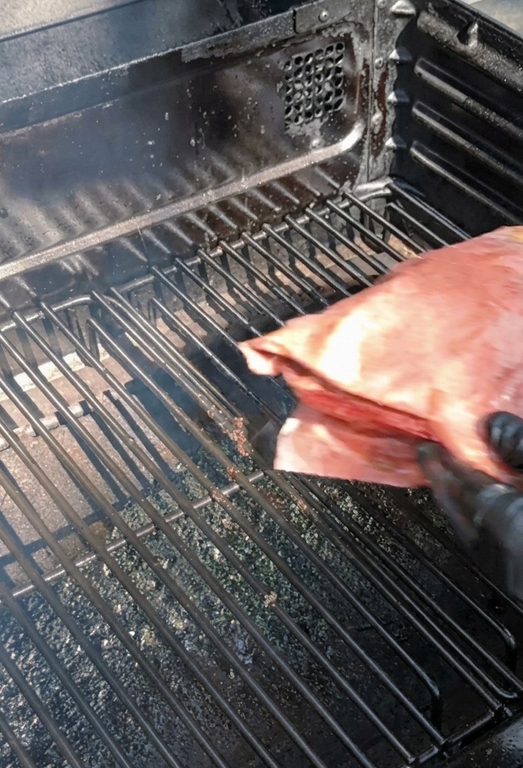 Eine Person in schwarzen Handschuhen legt ein großes, in rosa Butcher Paper eingewickeltes Paket auf den Grillrost eines Smokers.