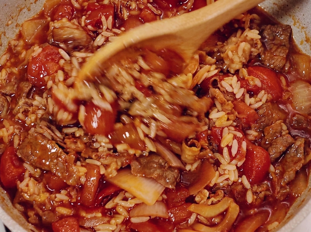 Ein Holzlöffel rührt Rindfleisch, Reis und Tomatensoße in der Pfanne um.