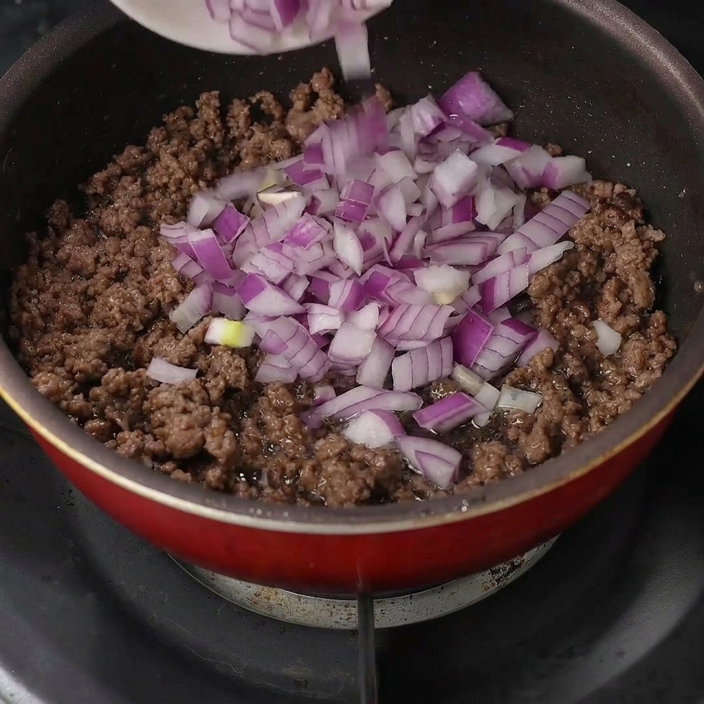 Frisch gewürfelte rote Zwiebeln werden über das gebratene Hackfleisch in der Pfanne gestreut.